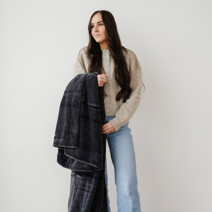 A woman holds a charcoal lush Saranoni blanket. This luxury blanket is a cozy blanket and a throw blanket. This soft blanket is great as a home decor staple, comfort gift, or everyday comfort item. Elevate your home and home decor with this faux fur blanket that is also a throw blanket. Enjoy this cozy blanket for years to come.