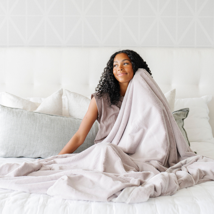 A woman holds a Feather (light gray) Lush XL Saranoni blanket, designed for oversized comfort and featuring a buttery soft, plush texture. This luxurious Saranoni blanket is perfect for cozy evenings, reading, or home decor. The Saranoni blanket adds warmth, style, and unmatched softness, making it an ideal gift or essential for ultimate relaxation.