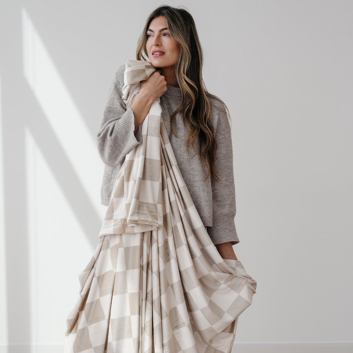 A woman holds a Saranoni luxury blanket that is a tan and cream checkered minky stretch blanket. Enjoy this soft blanket that is perfect for relaxing, gifting, or on the go.  Add this throw blanket to your collection!