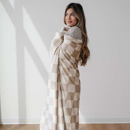 A woman is wrapped in a tan and cream checkered minky stretch blanket. This soft blanket is a Saranoni luxury blanket, and it is very stretchy, durable, and breathable. This extra large throw blanket is a very cozy blanket as well.